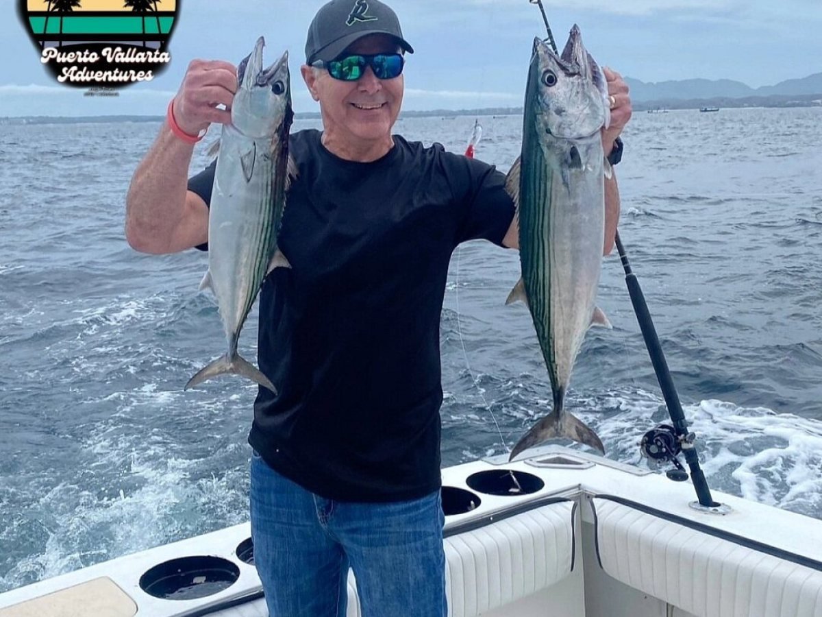 Smiling angler holding a bonito in each hand on the back deck of a charter boat in Banderas Bay