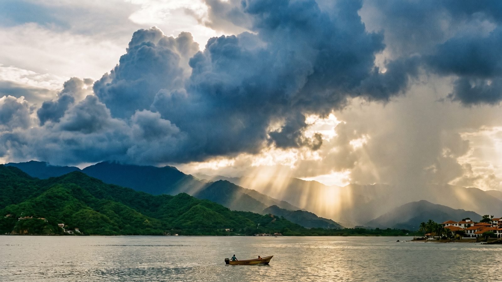 Storm clouds and sunbeams over Banderas Bay and the green Sierra Madre during Puerto Vallarta's rainy season, with a small panga on calm water